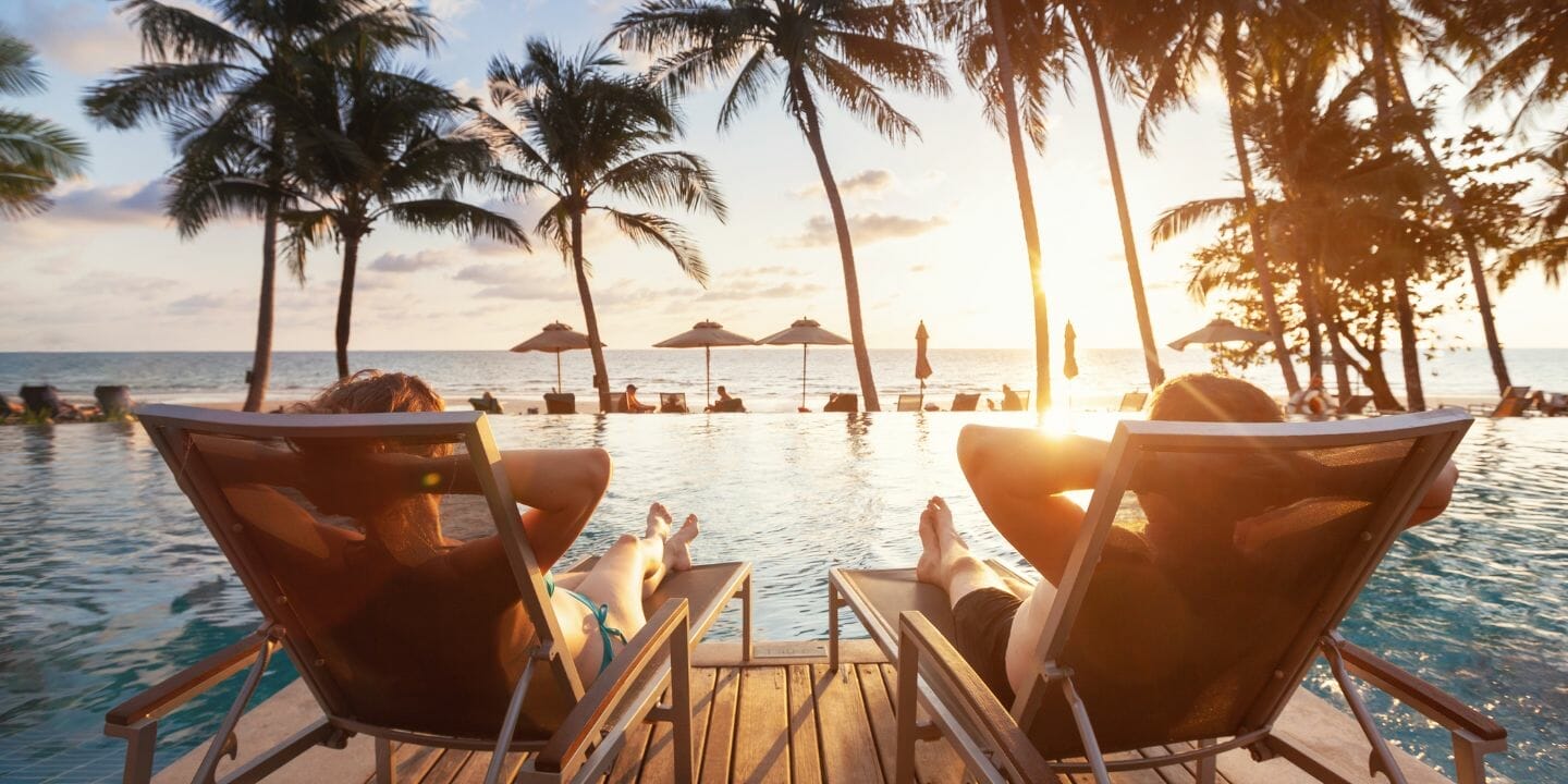 two people sitting in lounge chairs at the edge of a pool with palm trees in the background