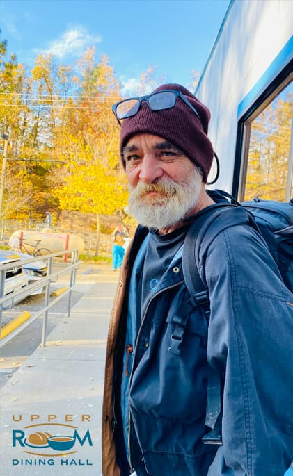 An elderly white bearded man in a windbreaker with hear gear cover and glasses. On the bottom right is the Upper Room Dining hall Logo