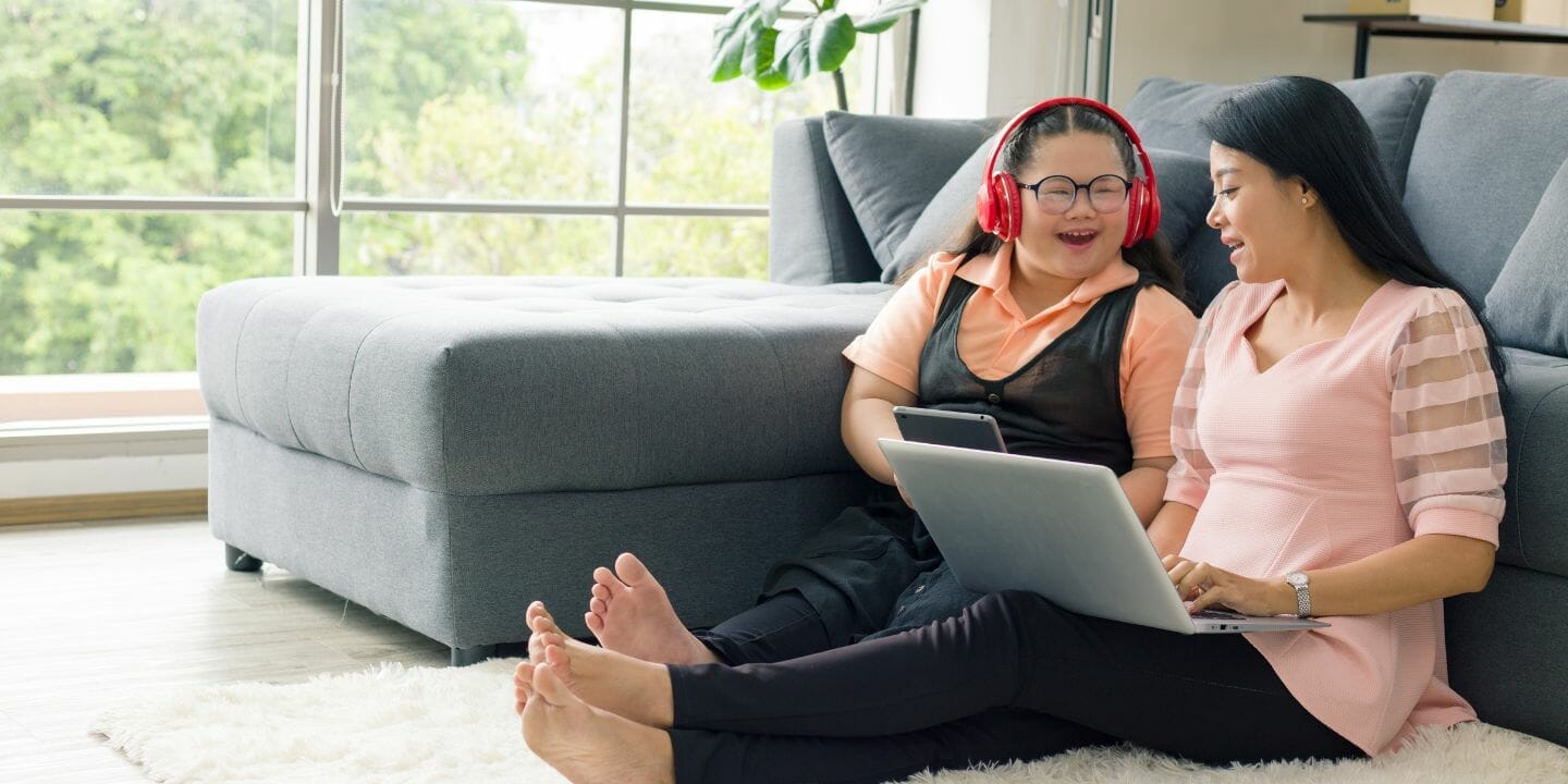 Two women sit on the floor by a gray sofa in a bright living room. One wears red headphones and holds a tablet, smiling. The other woman, in a pink top, is using a laptop and also smiling. Both appear to be enjoying their time together.