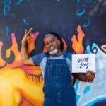 A Deaf Black man smiles and signs in American Sign Language while holding a "Deaf joy" print.