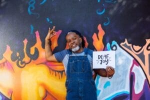 A Deaf Black man smiles and signs in American Sign Language while holding a "Deaf joy" print.