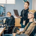 Man in wheelchair using assistive technology to use a computer. Woman with laptop stands beside him.