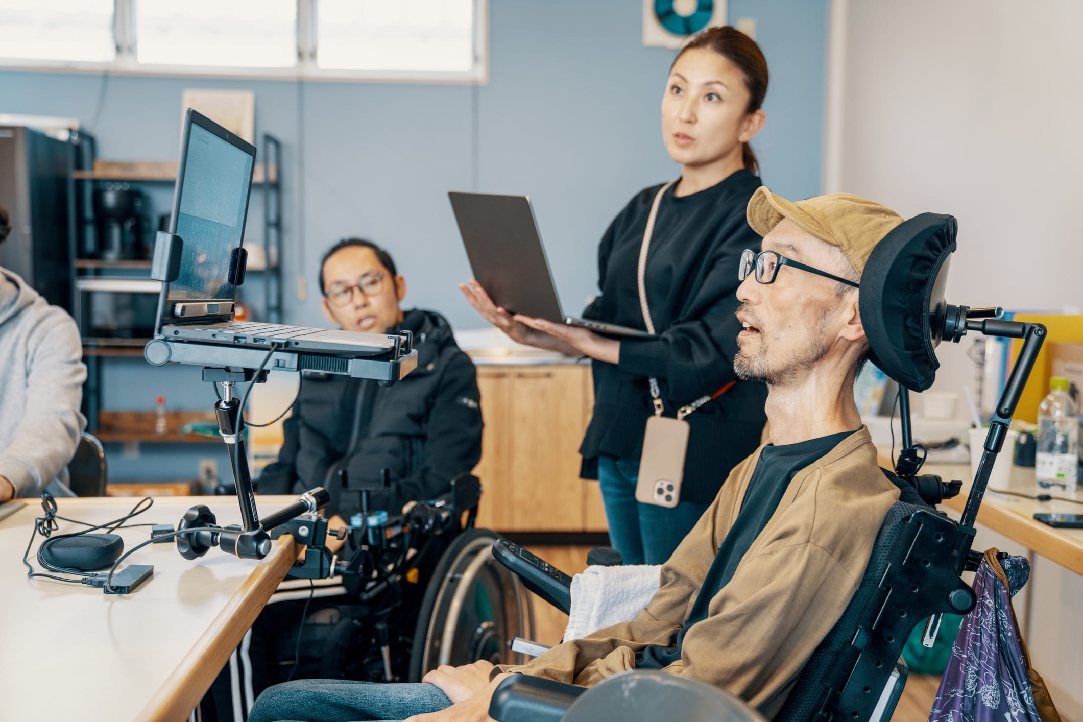 Man in wheelchair using assistive technology to use a computer. Woman with laptop stands beside him.