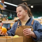 A woman with Down syndrome holding two oranges in her hands while checking a box of fruits.