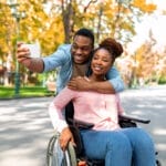 A couple takes a selfie outdoors, capturing an inclusive experience. The man stands behind and leans over the woman, who is sitting in a wheelchair amidst the autumn trees and park path—a perfect snapshot to inspire accessible gift ideas for cherished moments together.