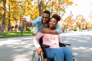 A couple takes a selfie outdoors, capturing an inclusive experience. The man stands behind and leans over the woman, who is sitting in a wheelchair amidst the autumn trees and park path—a perfect snapshot to inspire accessible gift ideas for cherished moments together.