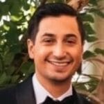 A man in a suit and bow tie stands smiling amidst leafy greenery,