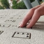 A hand reads raised braille dots on a textured surface, with greenery and blurred buildings visible in the background.