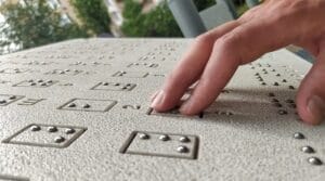 A hand reads raised braille dots on a textured surface, with greenery and blurred buildings visible in the background.