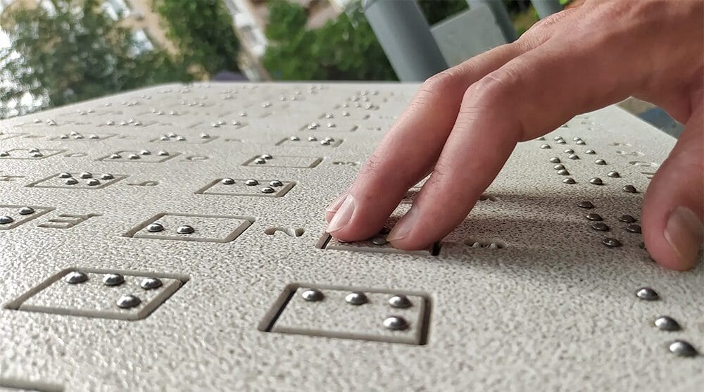 A hand reads raised braille dots on a textured surface, with greenery and blurred buildings visible in the background.