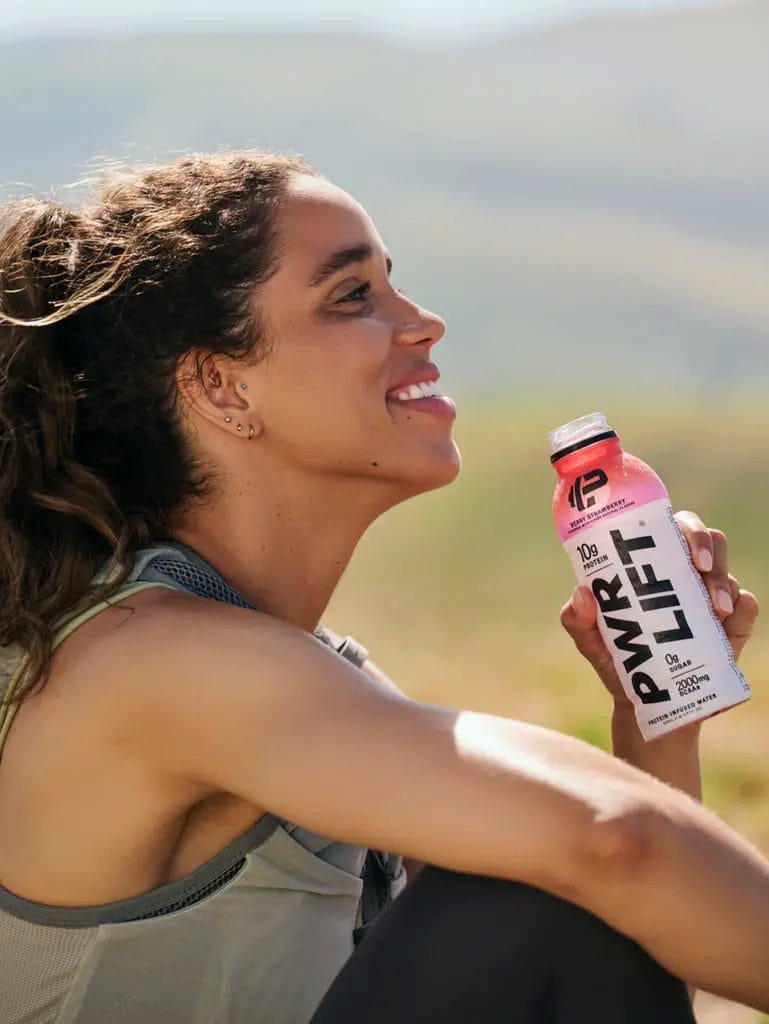 A woman sits outdoors, smiling and holding a bottle of PWR LIFT protein drink in her right hand.