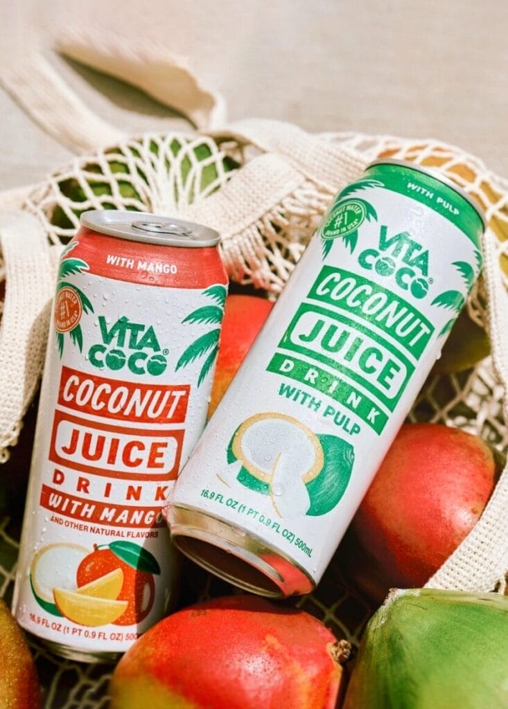 Two cans of Vita Coco Coconut Juice, one with mango and one with pulp, are placed in a mesh bag with mangoes and other fruits.