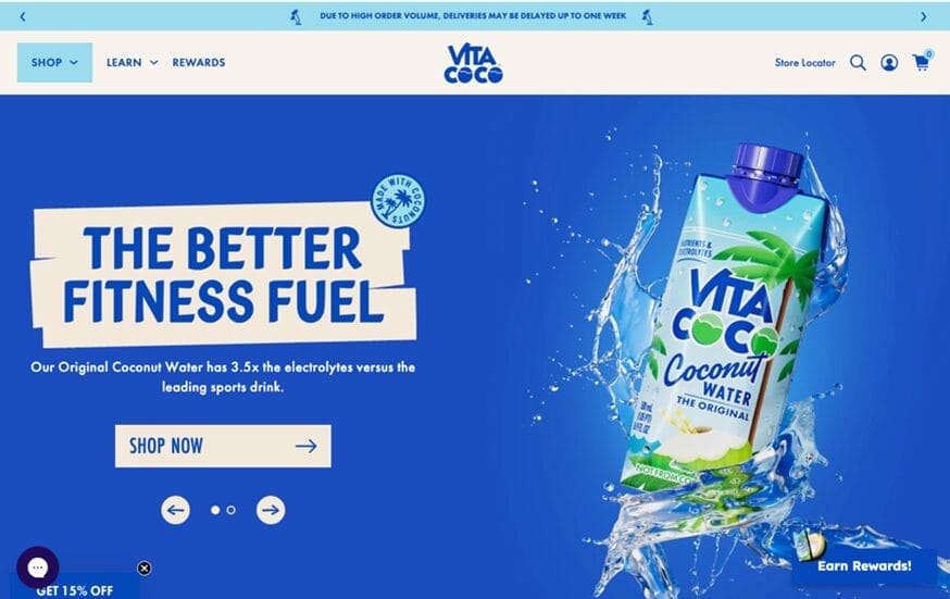 Vita Coco website homepage with a blue background. Left side shows “The Better Fitness Fuel” in bold white text on a blue paint streak. Right side features a splashing carton of Vita Coco Coconut Water with coconuts and palm trees on the packaging.